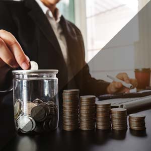 Business person placing coin into glass jar, next to stacks of other coins of varying heights