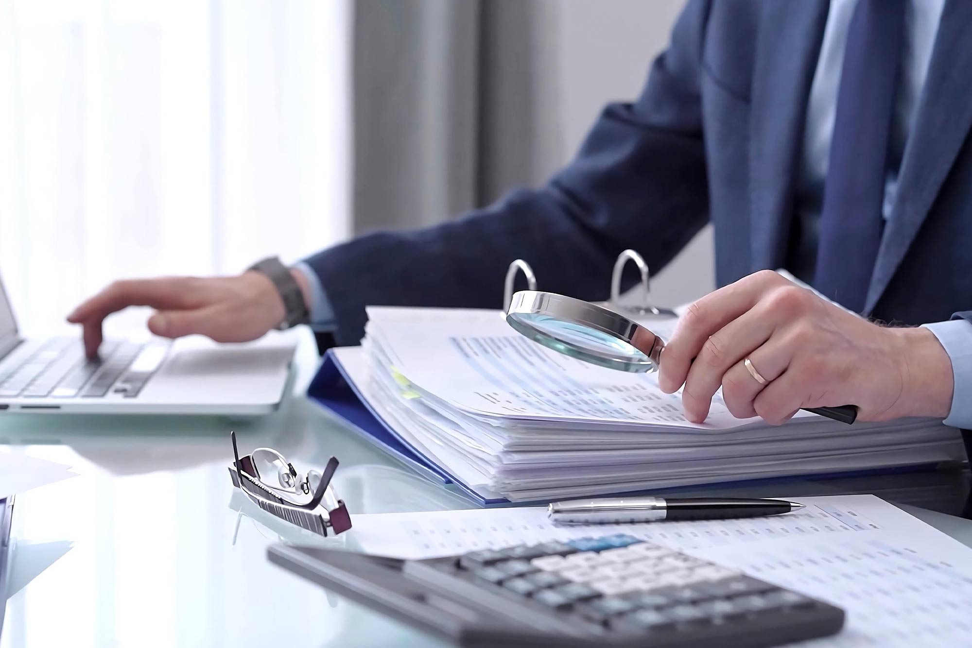 Businessman using a laptop computer and magnifying glass while analyzing financial documents at desk in modern office. Audit and taxes in business.