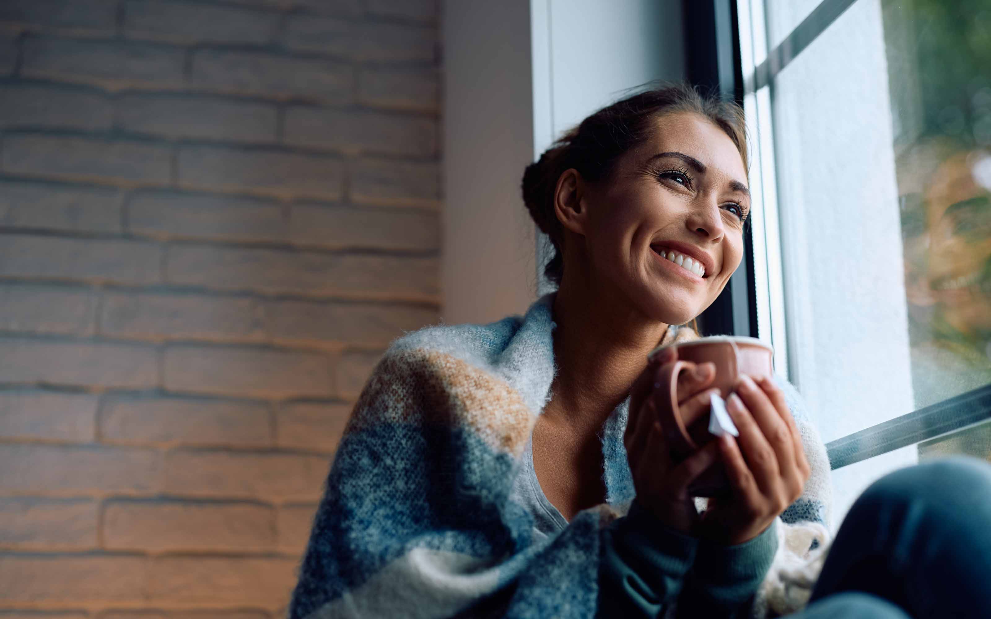 Smiling woman wrapped in a cozy blanket, enjoying a warm drink while sitting by an energy-efficient window.