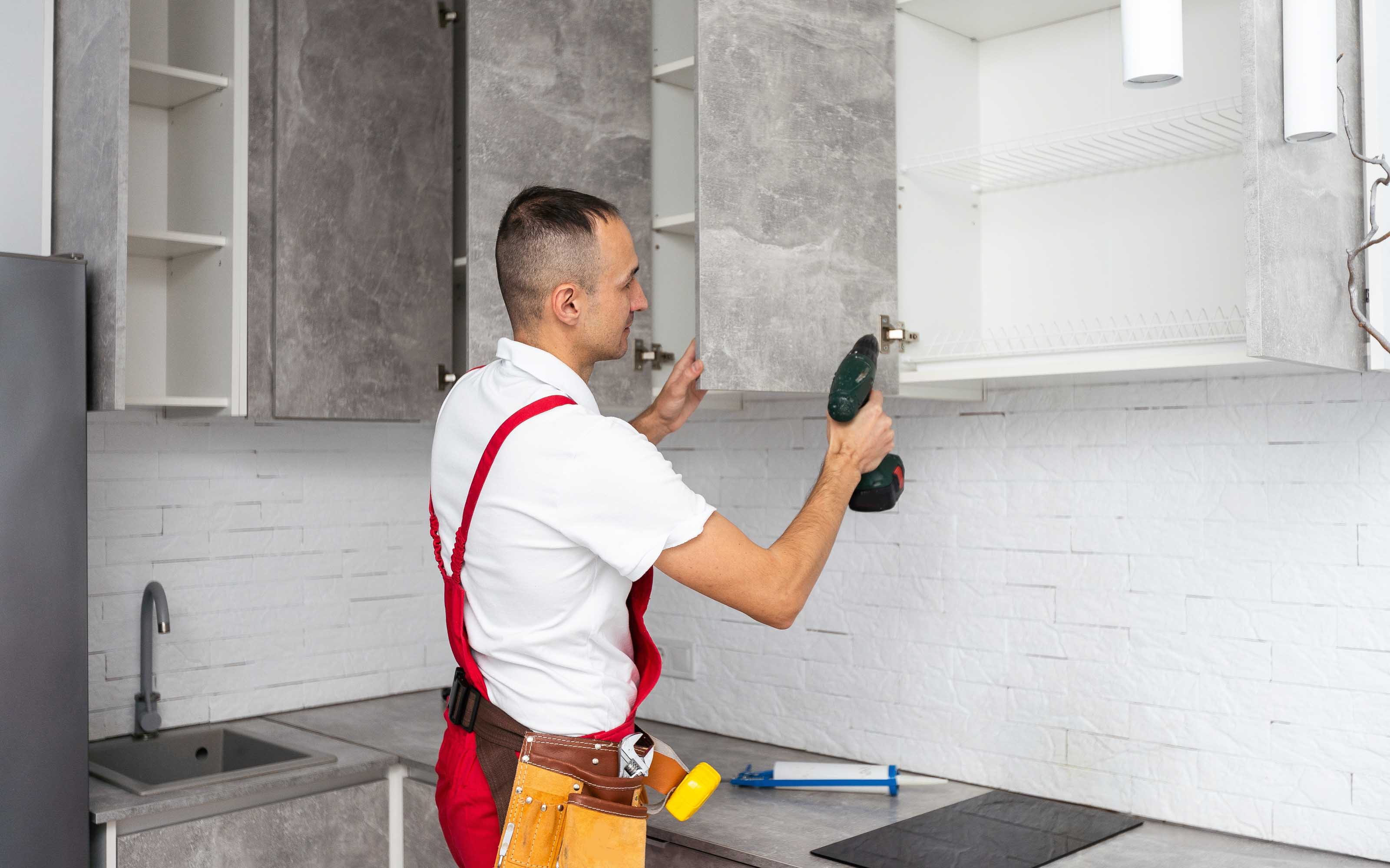 Kitchen Cabinet Installation in Progress Person installing a hinge on a gray upper kitchen cabinet using a green power drill, with tools on the counter in a modern kitchen setting.
