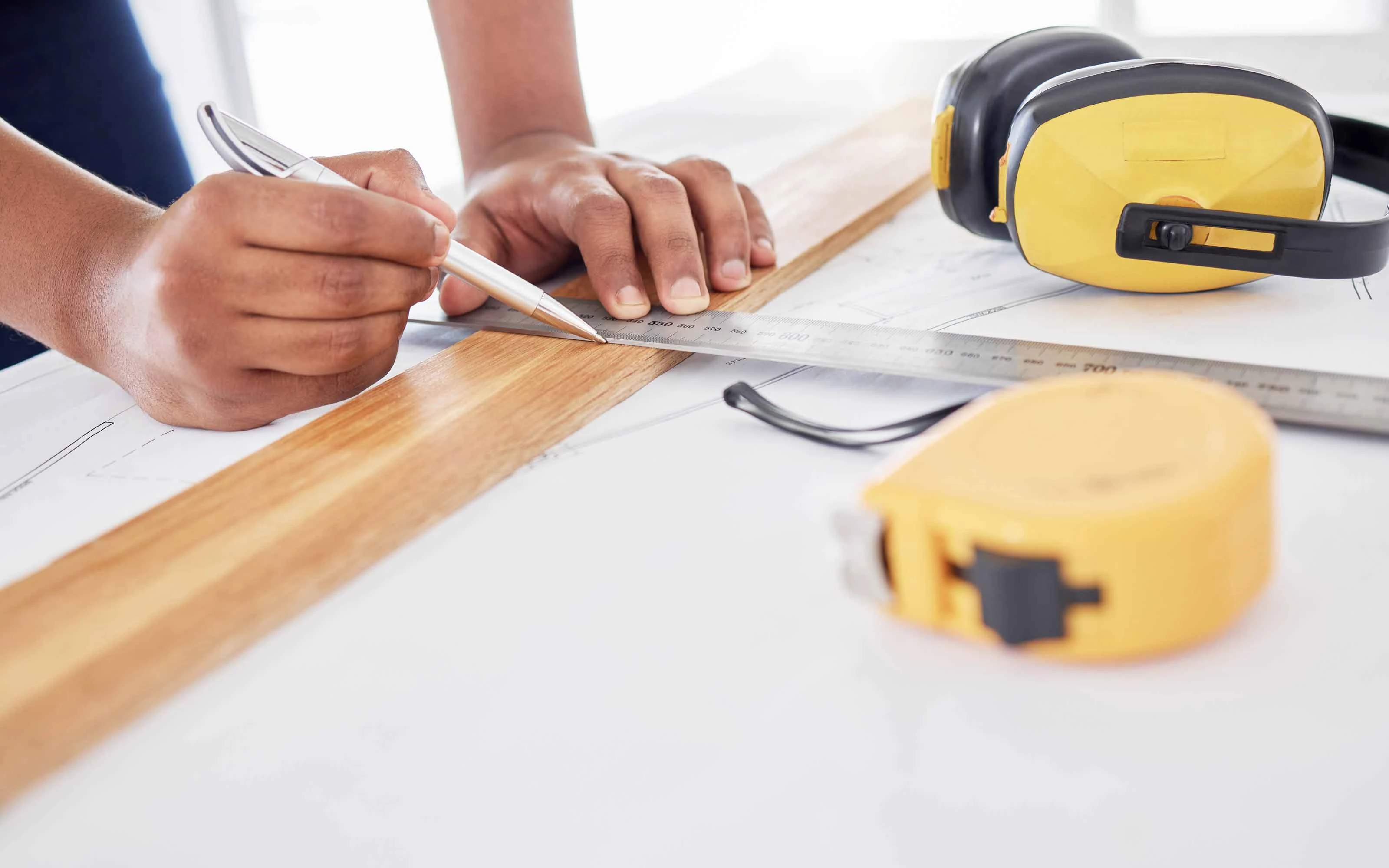 Person measuring a piece of wood with a ruler and pen, with ear protection headphones and a tape measure nearby