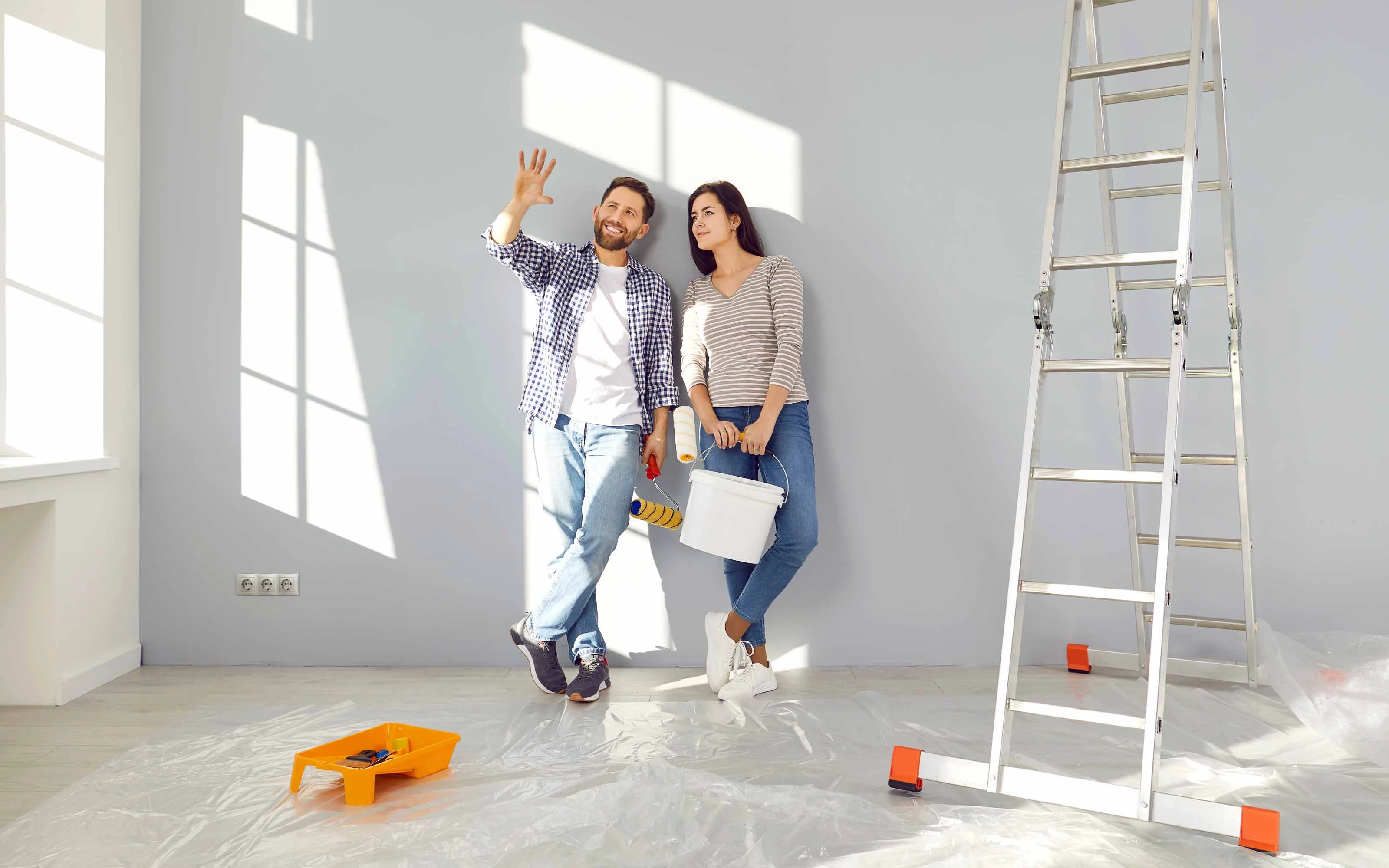 Two people in a room under renovation, with a paint roller, bucket, ladder, and tools on a plastic-covered floor