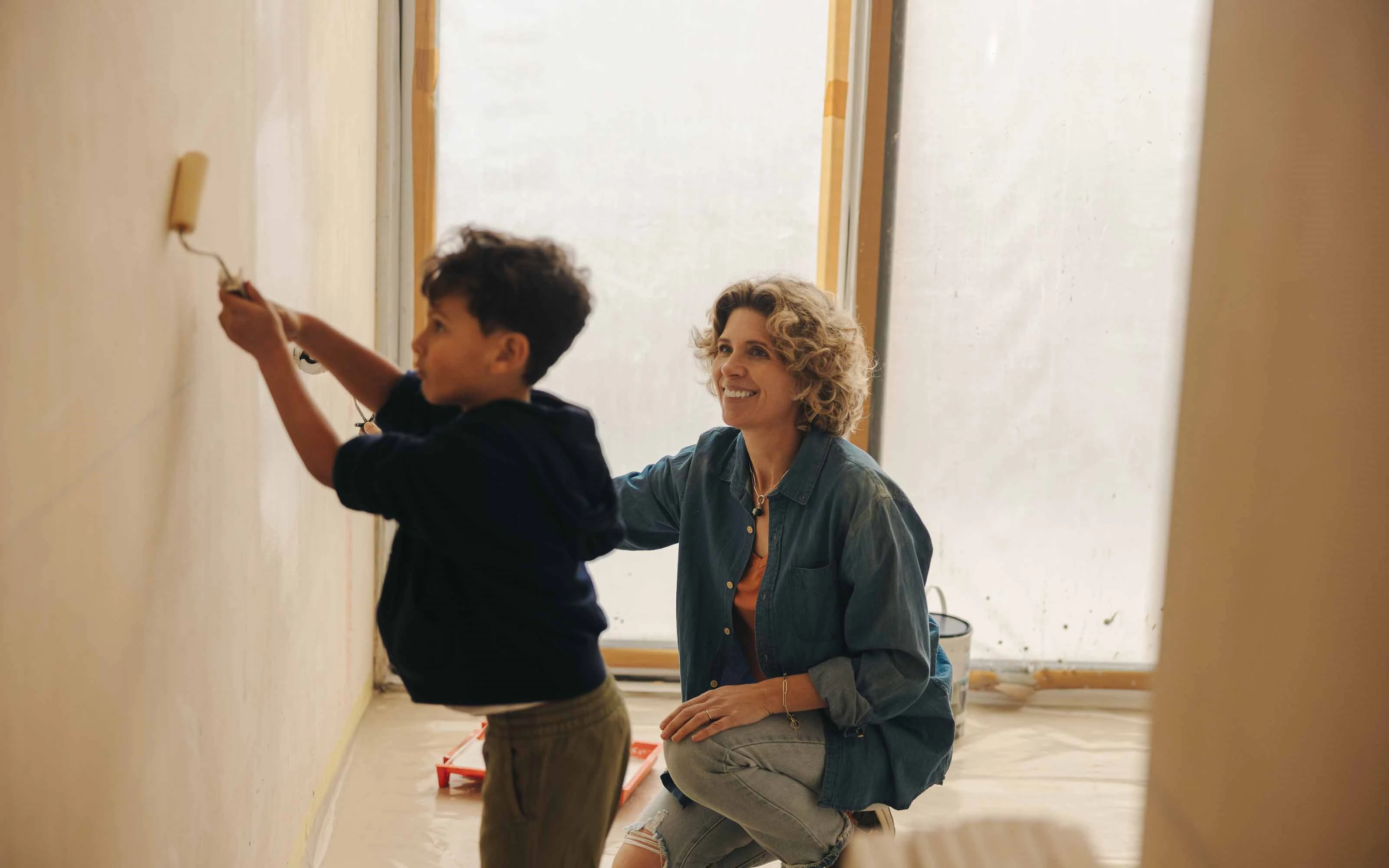 Child and adult painting a wall together in a room under renovation, with plastic sheets on the floor and windows
