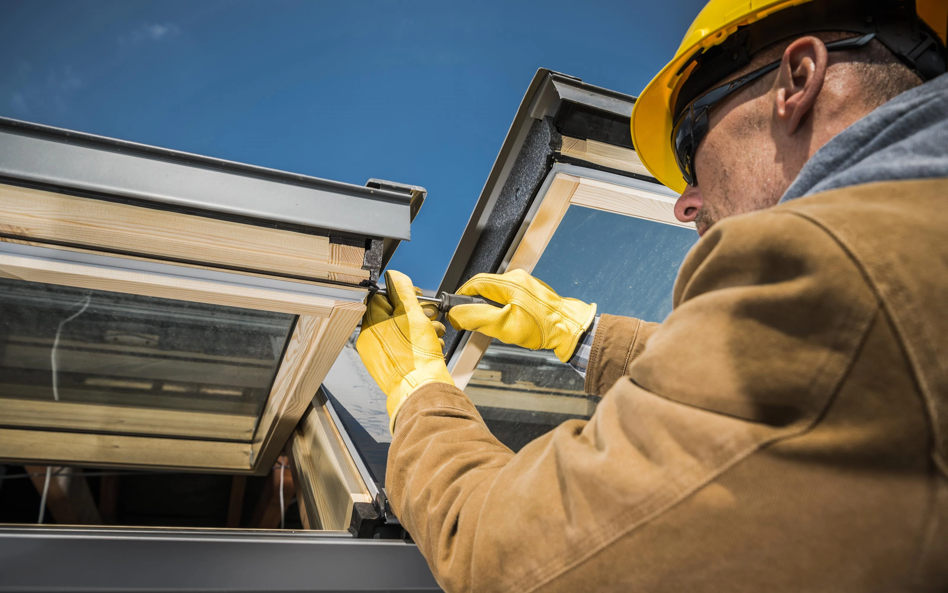 A worker wearing a yellow hard hat and gloves is installing or repairing a skylight window on a roof. The worker is using tools to secure the window frame.