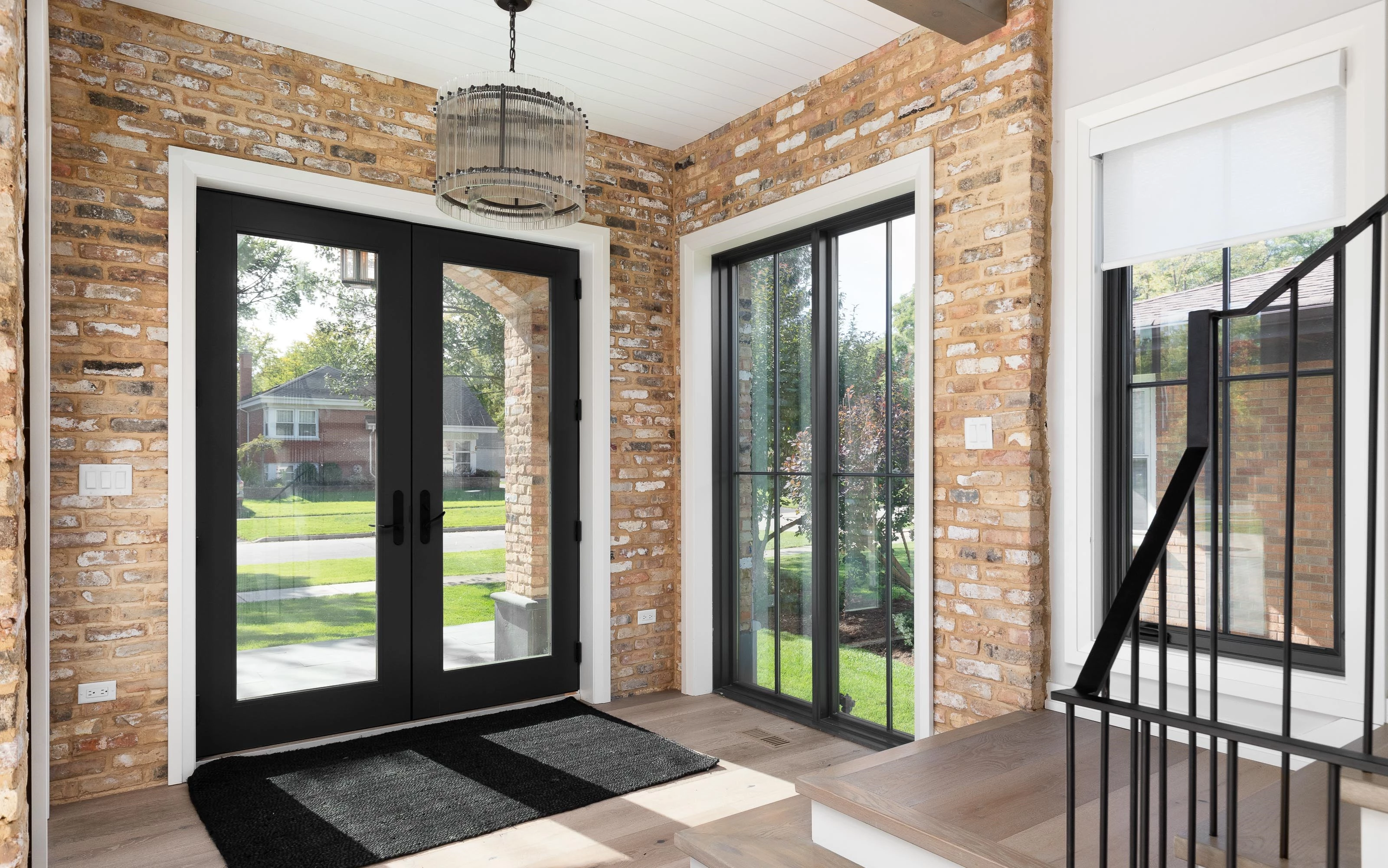 A bright and welcoming entryway with brick walls, large black-framed glass doors, and a modern chandelier.