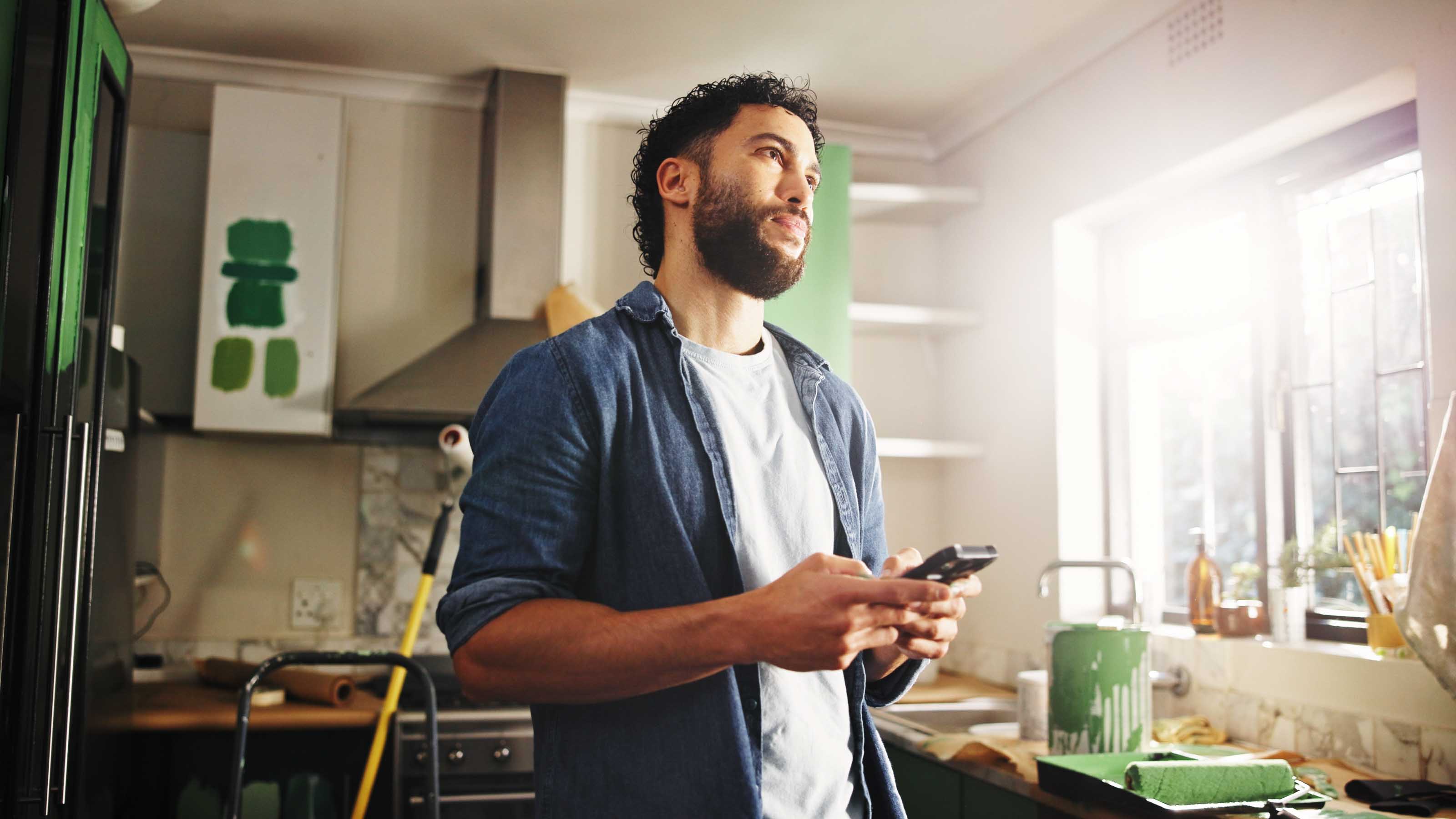 Man standing in a kitchen under renovation, holding a phone while painting supplies and green paint cans sit on the counter.