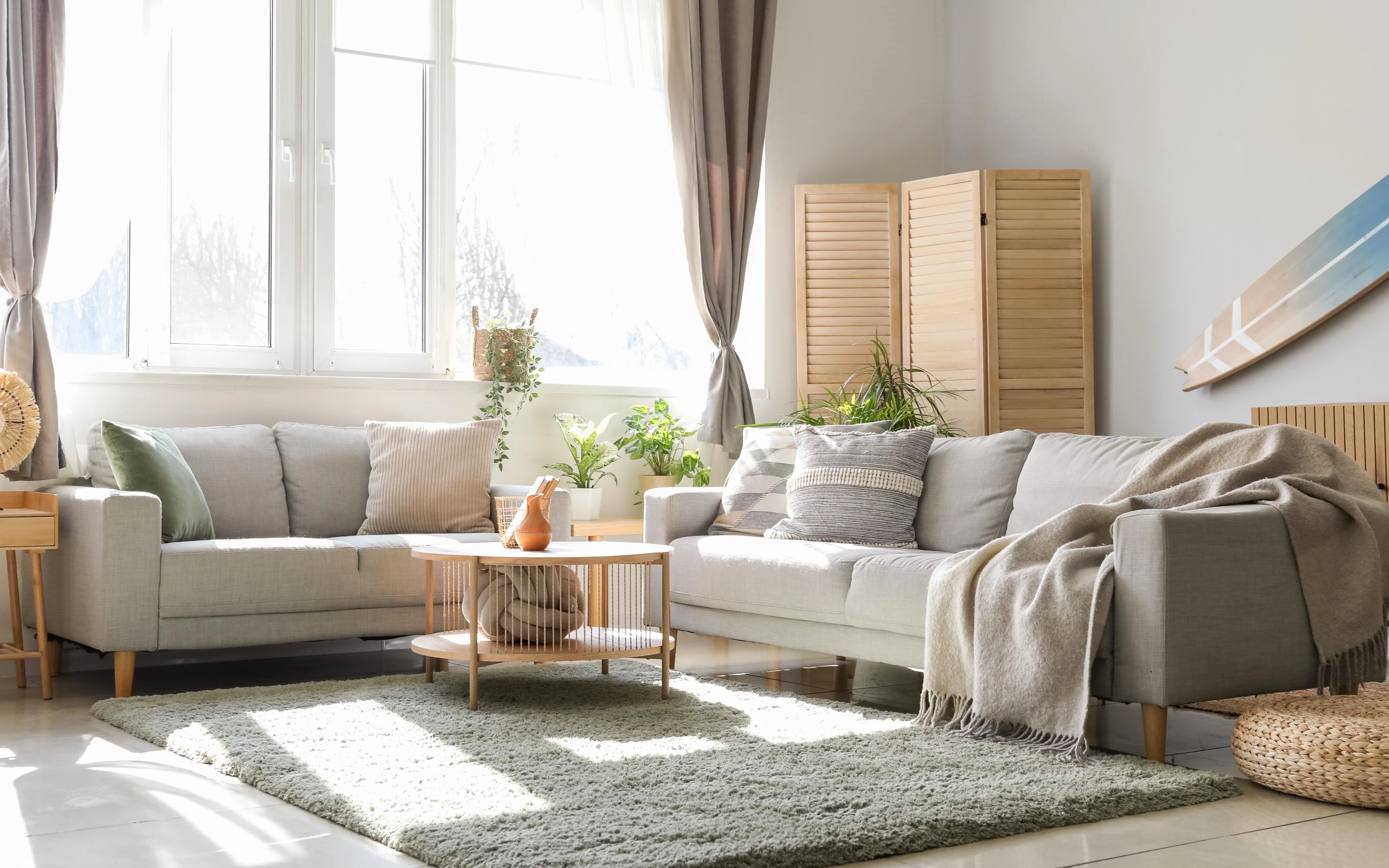 Bright and cozy living room with two light gray sofas, a wooden coffee table, and various plants. Sunlight streams through large windows with sheer curtains.