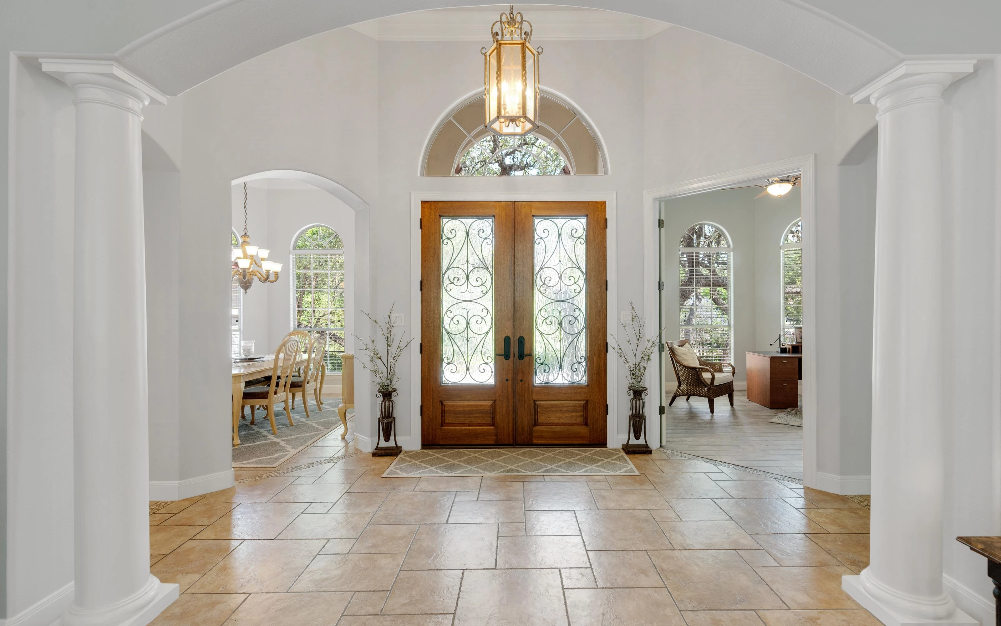 A spacious and elegant foyer with a grand wooden double door entrance, flanked by two white columns. The area is well-lit with natural light streaming through the arched windows and a chandelier hanging from the ceiling. The floor is tiled, and there are