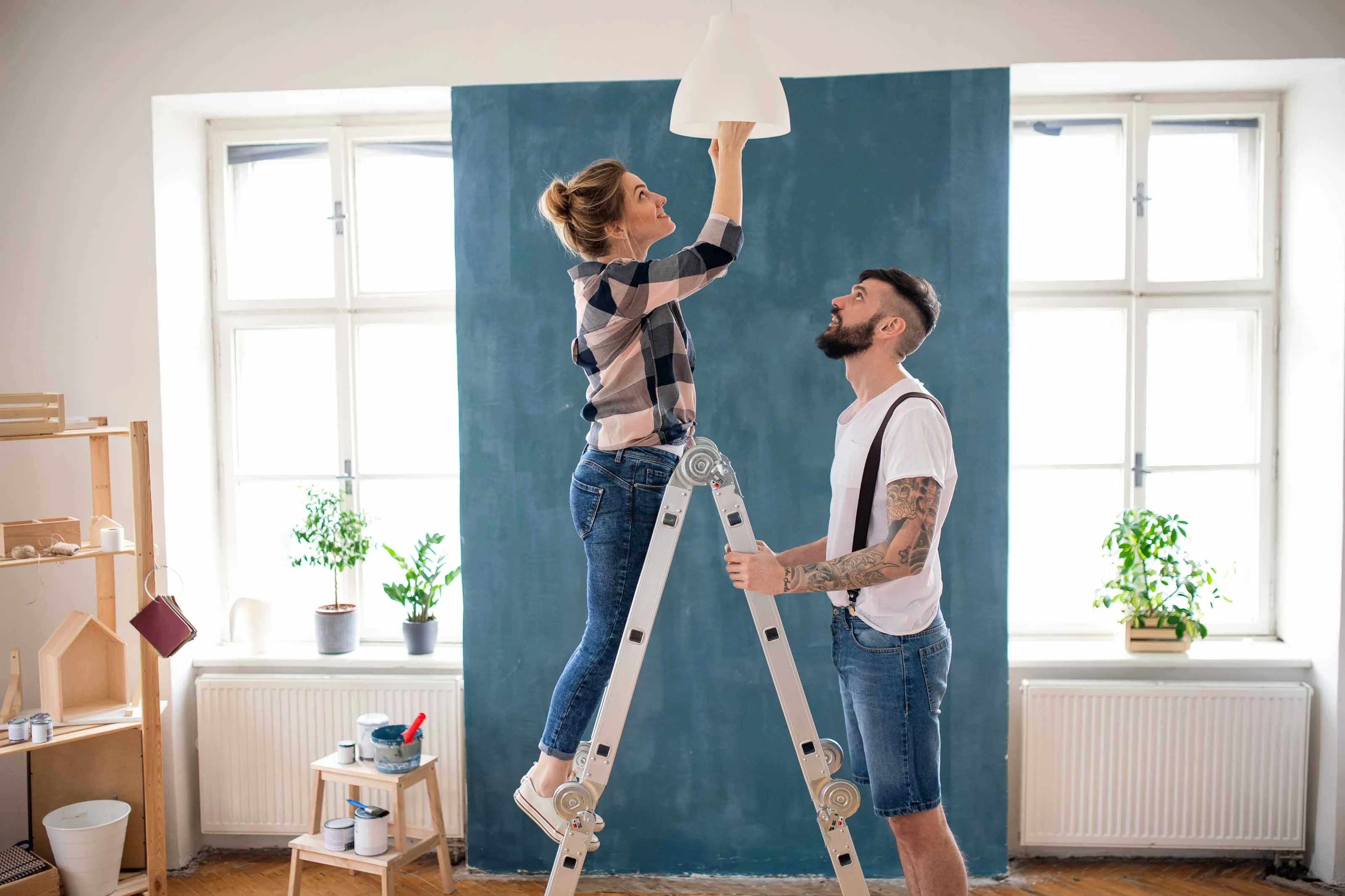 Woman Changing Light Fixture with Man Steadying Ladder A woman standing on a ladder changes a light fixture while a man steadies the ladder in a bright room with large windows and plants