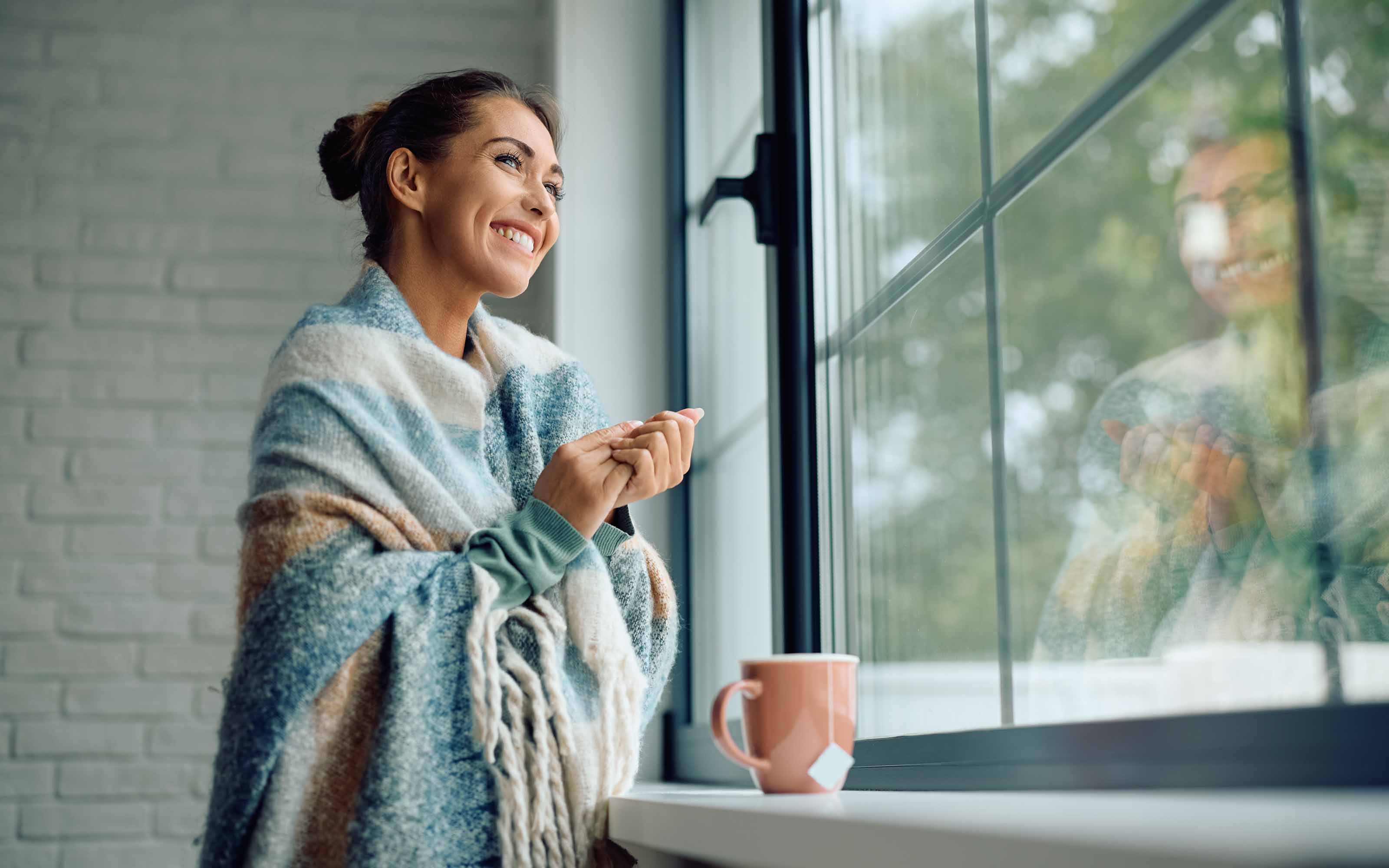 A smiling woman wrapped in a cozy blue and beige blanket looks out her window with a pink mug resting on the windowsill.