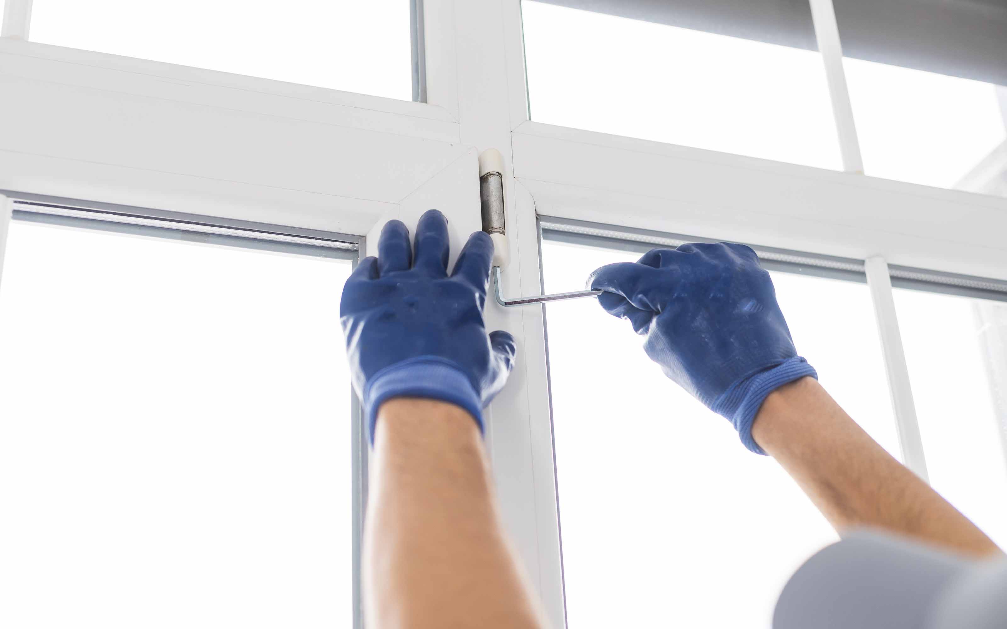 A worker wearing blue gloves uses a screwdriver to adjust the hinge on a white vinyl window frame.